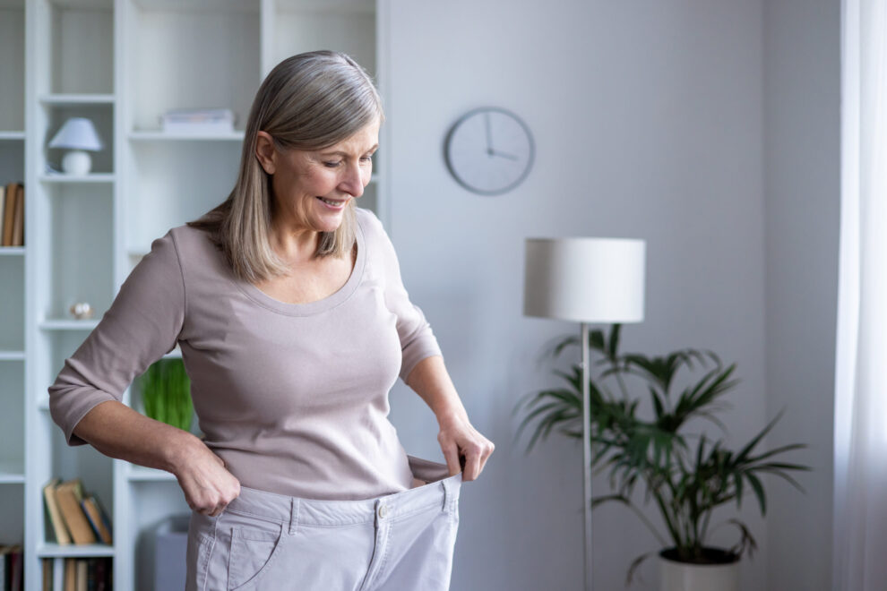 Happy senior woman examines her weight loss progress by wearing oversized pants. She stands in modern room, proud of healthy lifestyle and personal achievement in fitness journey.